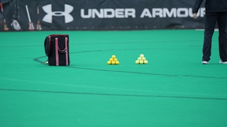 A sports field features a black and pink sports bag, two pyramids of yellow balls, and a portion of a person in dark athletic gear. The background displays a banner with the Under Armour logo.
