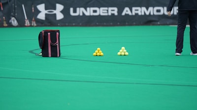 A sports field features a black and pink sports bag, two pyramids of yellow balls, and a portion of a person in dark athletic gear. The background displays a banner with the Under Armour logo.