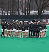 A group of athletes is gathered in a huddle on a sports field, wearing matching uniforms and gear. The ground is green, and there is signage with a brand logo in the background. Trees and parked cars can be seen behind a fence surrounding the field. A person on the left side is capturing the moment with a camera.