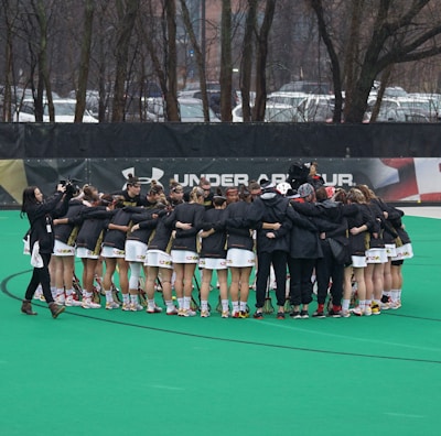 A group of athletes is gathered in a huddle on a sports field, wearing matching uniforms and gear. The ground is green, and there is signage with a brand logo in the background. Trees and parked cars can be seen behind a fence surrounding the field. A person on the left side is capturing the moment with a camera.