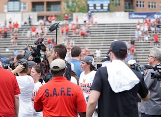 A lively gathering in an outdoor sports venue with a group of people, some wearing matching shirts labeled 'National Champions.' Several individuals are holding cameras and microphones, indicating a media presence. The background shows blurred spectators sitting on bleachers.
