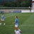 A soccer game scene on a green grass field with players wearing light blue jerseys and white shorts. A goalpost is visible in the background, and there are people sitting or standing on the bleachers. Advertising banners are displayed along the sidelines.