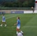 A soccer game scene on a green grass field with players wearing light blue jerseys and white shorts. A goalpost is visible in the background, and there are people sitting or standing on the bleachers. Advertising banners are displayed along the sidelines.