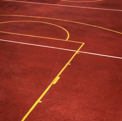 A sports court with a reddish-brown surface and bright yellow and white lines. The lines form geometric shapes, including rectangles and circles, suggesting markings for games like basketball or netball.