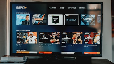 A television screen displaying the ESPN+ streaming interface with various sports-related shows and categories. The screen includes sections such as 'Browse by Shows' and 'Best of Drew Brees' with thumbnails of sports programs featuring athletes in action and analysts.