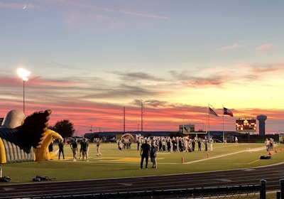 A sports field with a football game unfolding under a vibrant sunset sky, featuring a large inflatable mascot and a scoreboard displaying the score. Players and coaches are visible on the green field, surrounded by a track and some flags waving in the breeze.