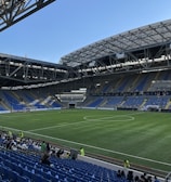 A modern sports stadium with a large, green soccer field and surrounding seating areas. Blue and yellow seats are visible, with some spectators scattered across the stands. The stadium is partially covered with a steel lattice roof, showing a clear blue sky. Several security personnel in high-visibility vests are present.