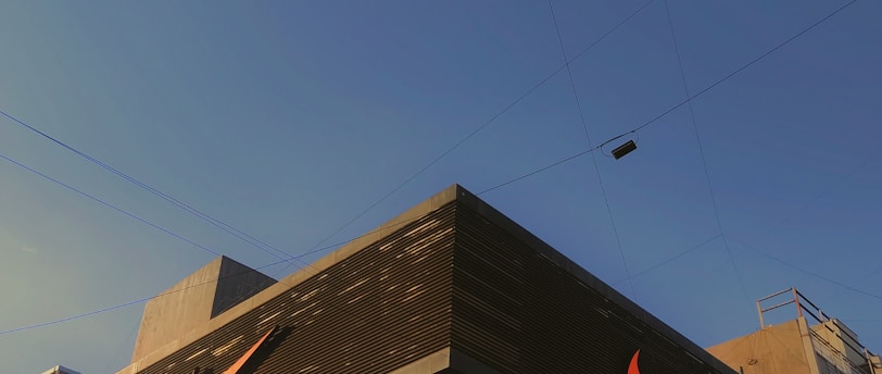 A modern building with predominantly gray and black tones featuring a large, well-known sports logo in bright orange. The facade includes glass panels displaying large advertisements. The sky is clear and blue, suggesting a sunny day. Nearby utility poles and wires are also visible.