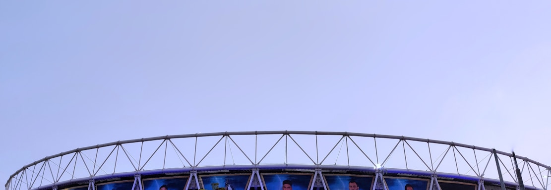 A large stadium with a circular design features multiple large banners displaying images of sports players in blue uniforms. The structure is modern, with a metal frame and triangular patterns. A crowd of people is gathered below, walking toward the entrance under a clear sky.