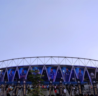 A large stadium with a circular design features multiple large banners displaying images of sports players in blue uniforms. The structure is modern, with a metal frame and triangular patterns. A crowd of people is gathered below, walking toward the entrance under a clear sky.