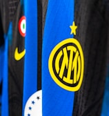 Close-up view of a sports jersey featuring bold vertical stripes in blue and black, adorned with a prominent yellow logo featuring a star on top. Additional elements include a Nike swoosh in yellow, a circular emblem with red, white, and green colors, and a section with black stars on white.