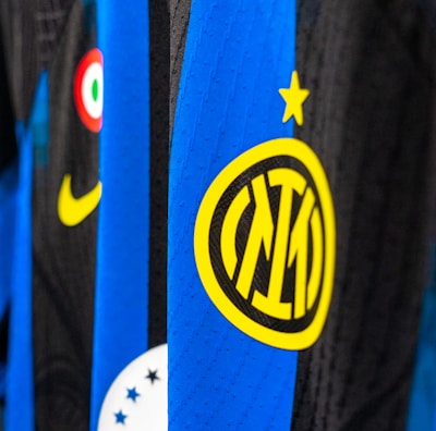 Close-up view of a sports jersey featuring bold vertical stripes in blue and black, adorned with a prominent yellow logo featuring a star on top. Additional elements include a Nike swoosh in yellow, a circular emblem with red, white, and green colors, and a section with black stars on white.