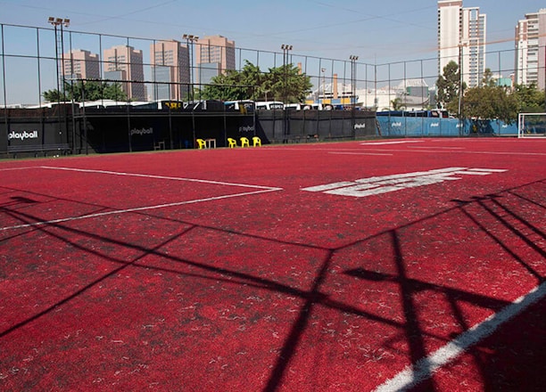 A sports court with a red playing surface and white lines, surrounded by a black mesh fence. In the background, tall buildings are visible, along with some trees and a few yellow chairs positioned near the fence.