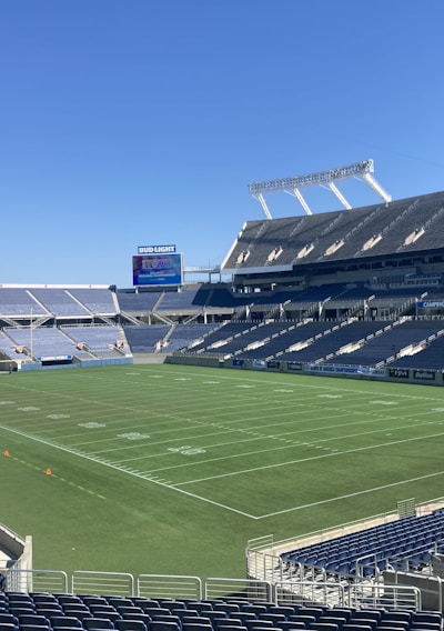 A large, empty sports stadium with blue seats surrounding a well-maintained green grass field. Bright sunlight creates clear shadows, and there are two goal posts on opposite ends of the field. A big electronic scoreboard displays some graphics, and the sky is clear and blue.